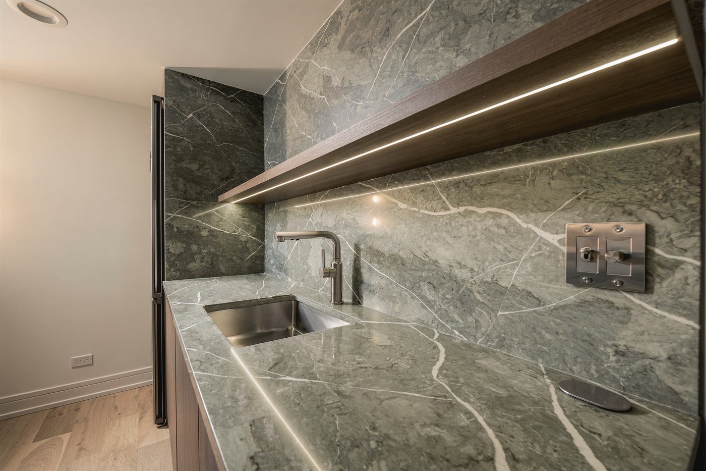 Green marble counter and backsplash with undermount sink beneath the lit walnut shelf